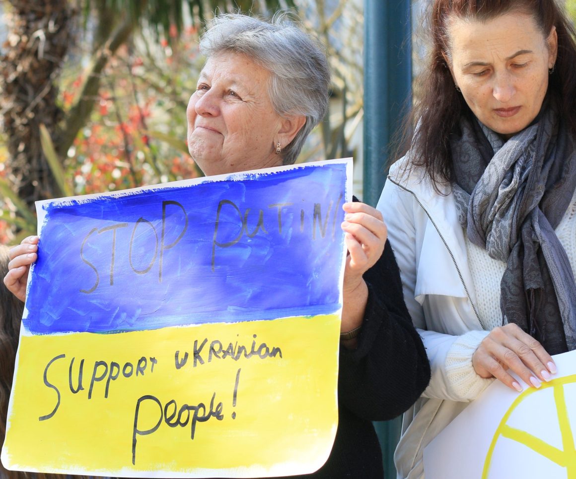 An elderly woman holds a placard that reads, 'Stop Putin, support Ukrainian people'