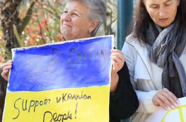 An elderly woman holds a placard that reads, 'Stop Putin, support Ukrainian people'