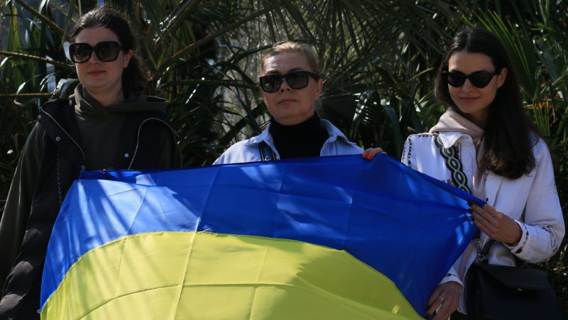 Three women wearing dark sunglasses and holding a Ukrainian flag