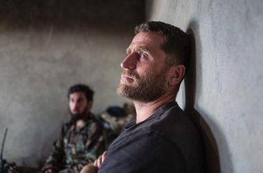 In the foreground there is a War Journalist, male, in a war zone with a soldier, male, in the background.
