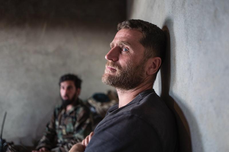 In the foreground there is a War Journalist, male, in a war zone with a soldier, male, in the background.