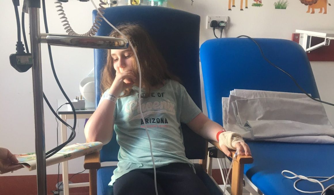 Photo of a young girl in a hospital chair wired up to a machine as she is very unwell.