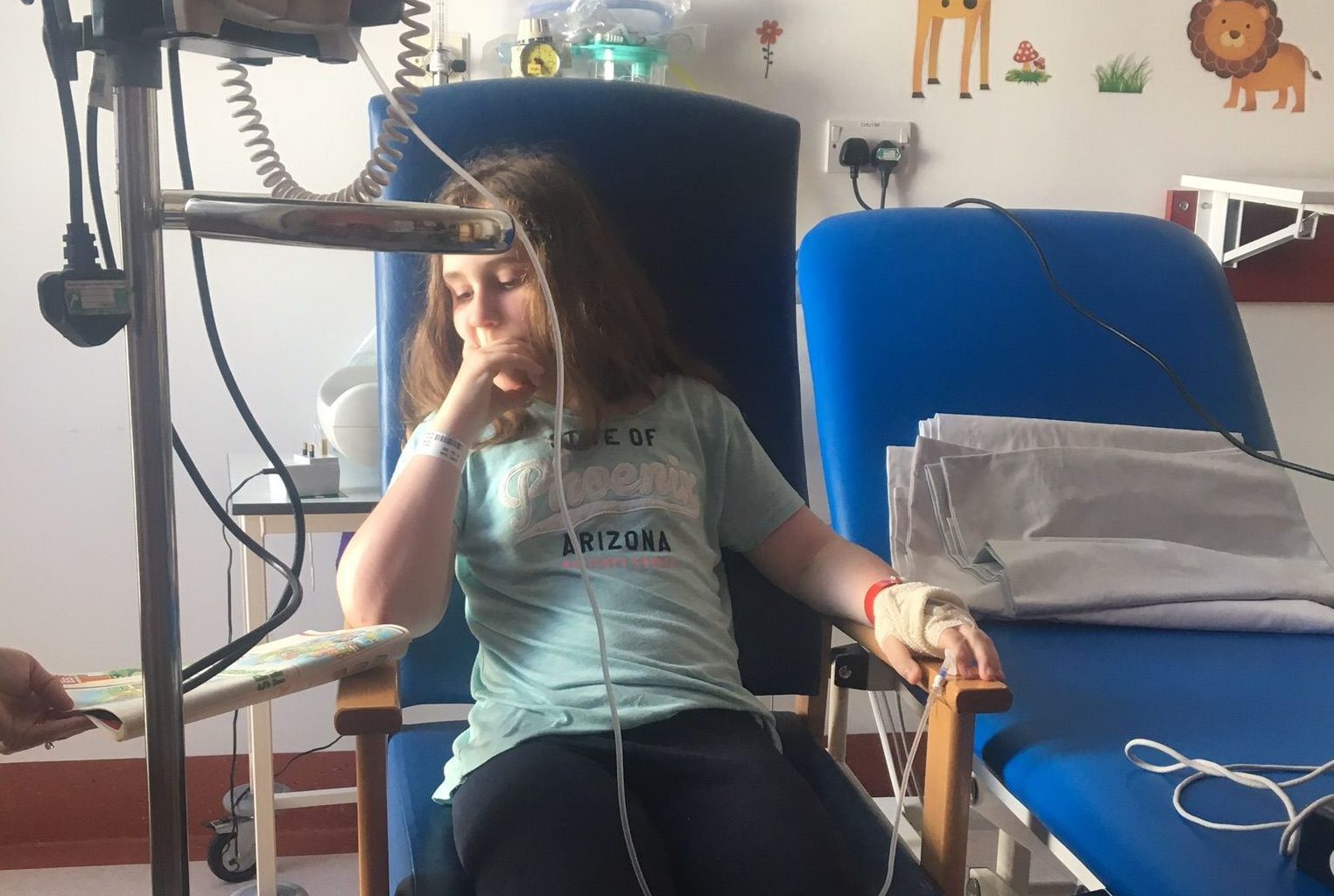 Photo of a young girl in a hospital chair wired up to a machine as she is very unwell.