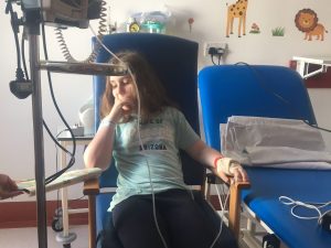 Photo of a young girl in a hospital chair wired up to a machine as she is very unwell.