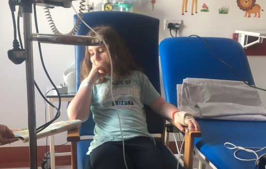 Photo of a young girl in a hospital chair wired up to a machine as she is very unwell.